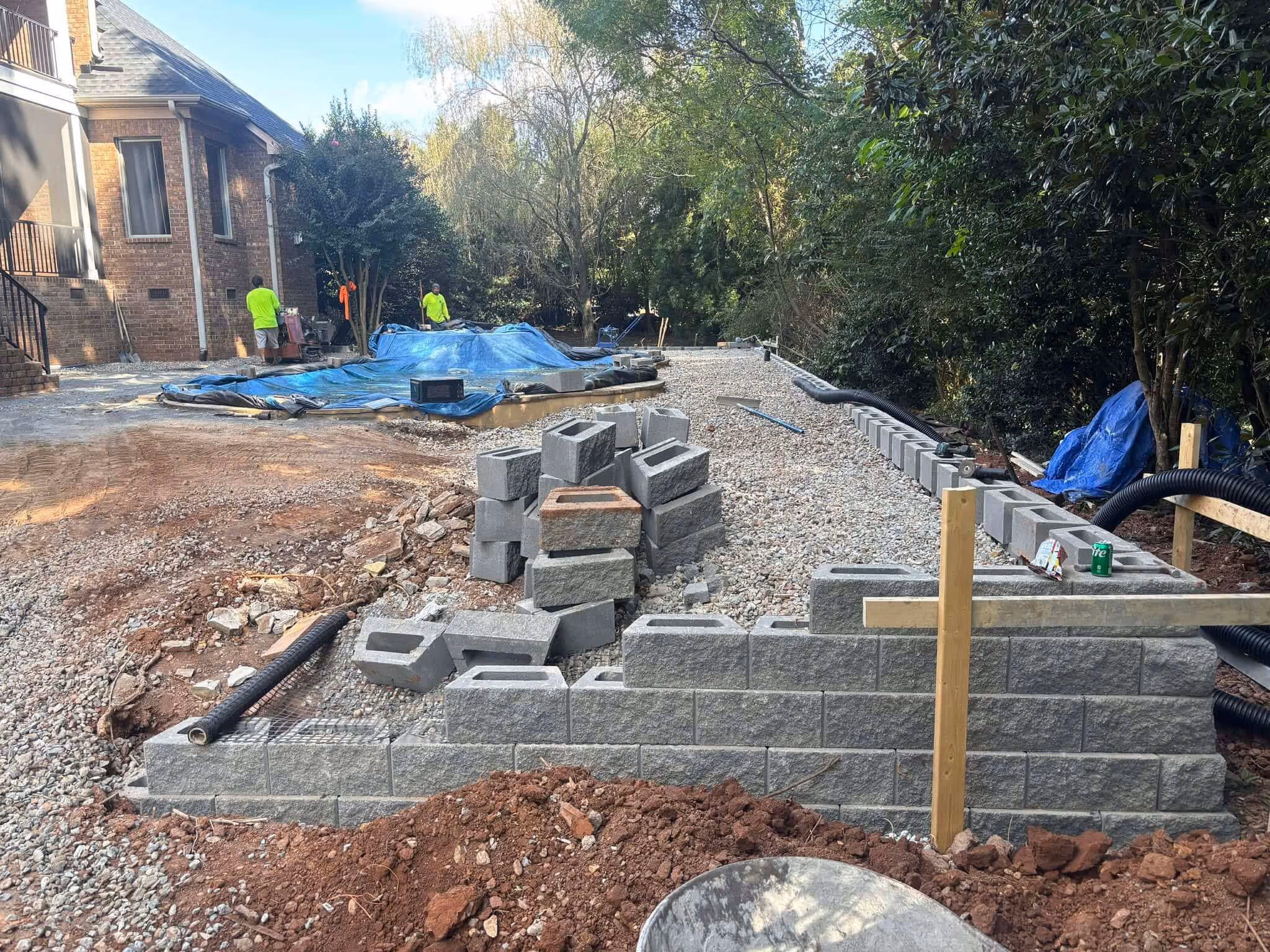 Construction site with workers assembling a gray concrete block retaining wall beside a house, surrounded by trees and gravel.