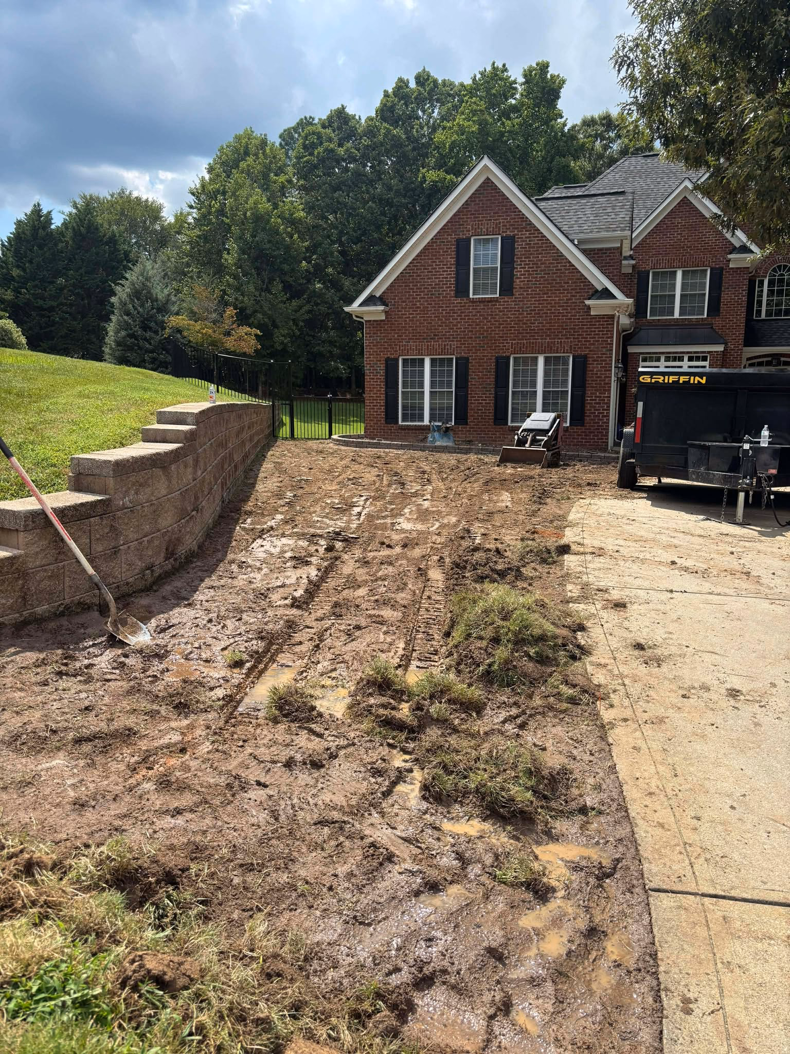 Rutted, muddy front yard beside a retaining wall with the lawn stripped back to bare soil.