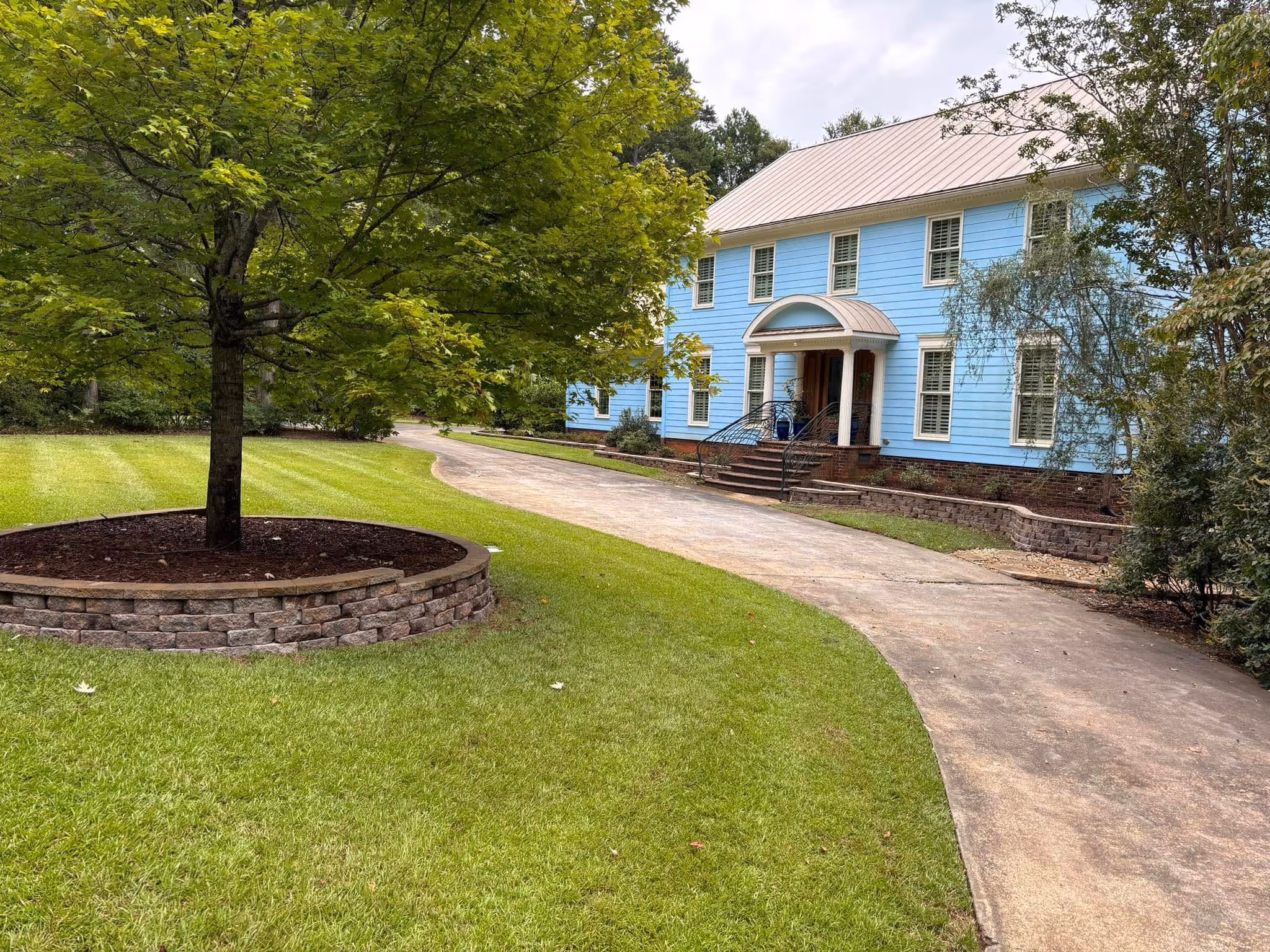 Wide mown lawn running up to a two-story home with a curved driveway and mature shade trees.