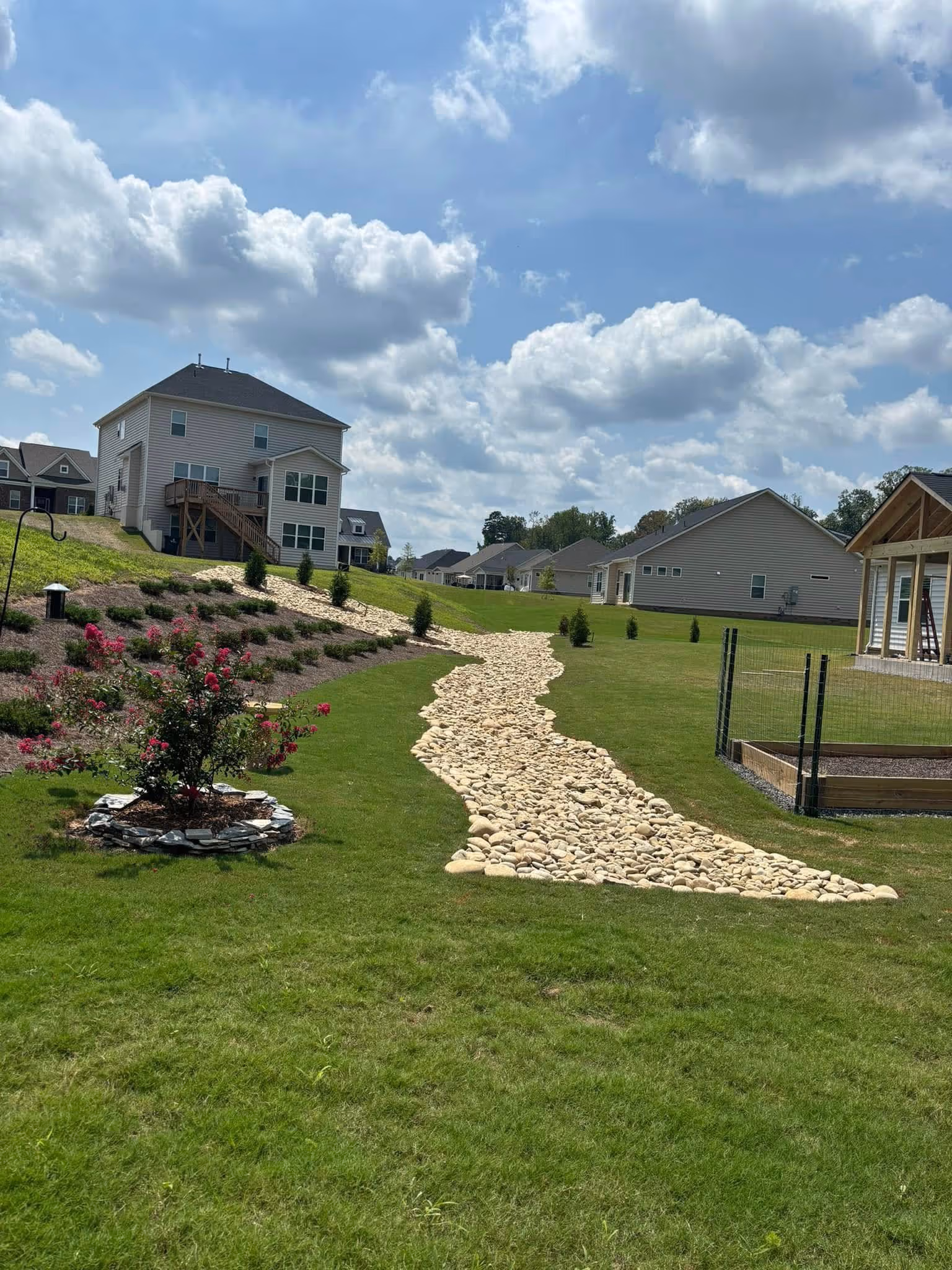 Residential backyard with a winding dry creek bed made of smooth white stones, green grass, rose bushes, and houses under a partly cloudy sky.