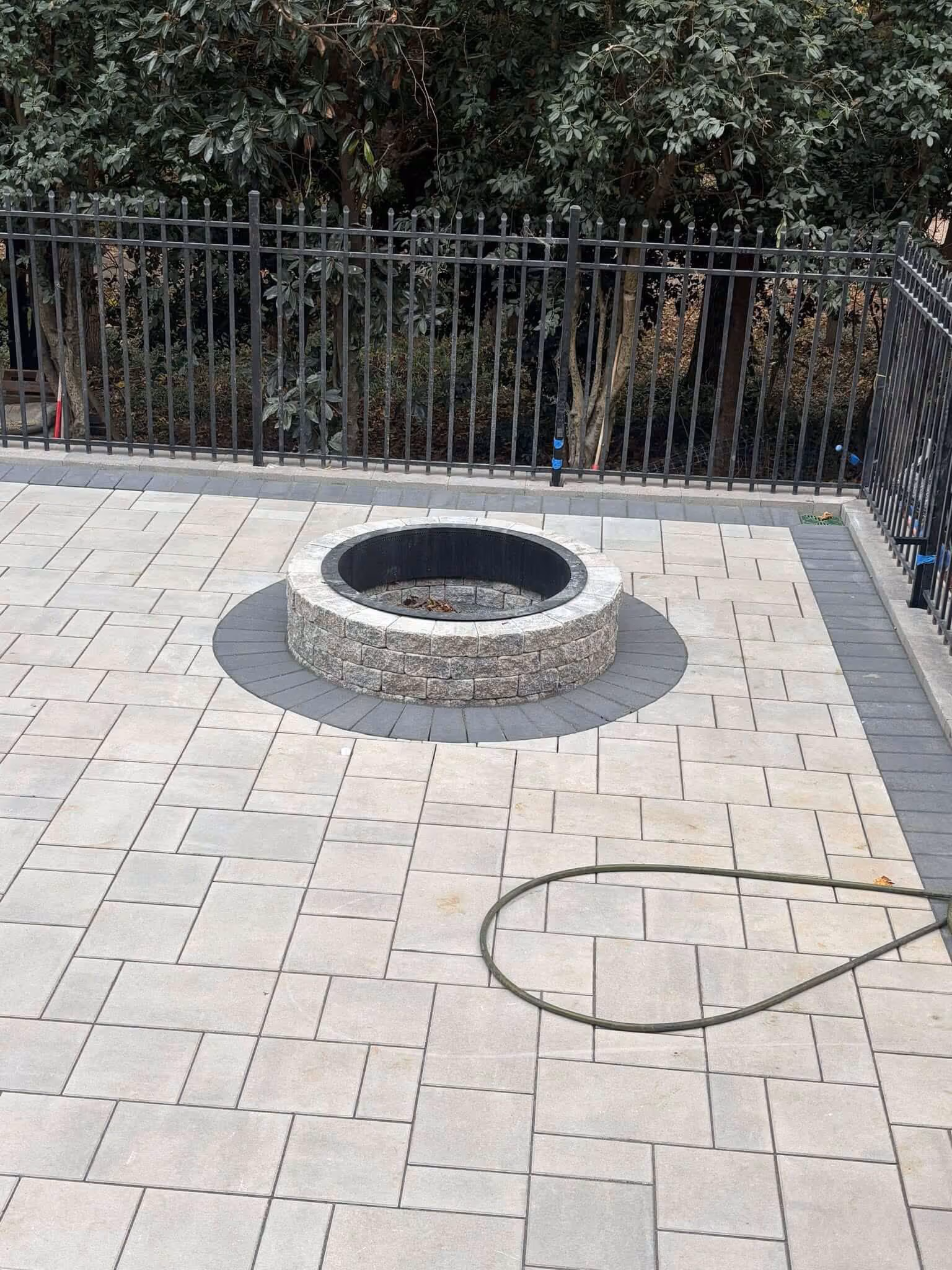 Circular stone fire pit surrounded by light-colored patio pavers with a coiled garden hose on the ground and a black metal fence with trees in the background.