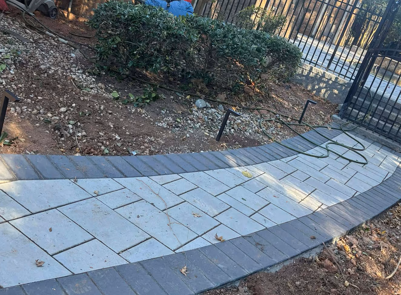 Curved paved walkway with light and dark gray bricks leading to a black metal garden gate surrounded by bare soil and bushes.